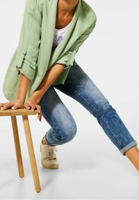 Jasnozielona, luźna marynarka noszona na białej koszuli, zestawiona z wyblakłymi niebieskimi dżinsami podwiniętymi u dołu. Beżowe trampki, drewniany stołek.