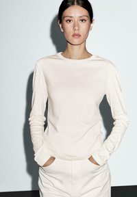 Crème kleurige longsleeve top van zachte, rekbare stof met een ronde halslijn; gedragen met de handen in de zakken, gecombineerd met bijpassende onderkleding.