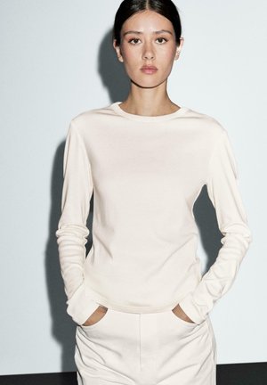 Cremfarbener Longsleeve in weichem, dehnbarem Stoff mit Rundhalsausschnitt; wird mit den Händen in den Taschen getragen und mit passenden Hosen kombiniert.