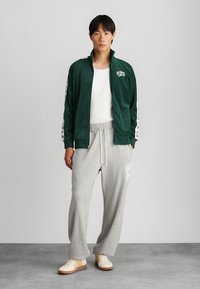 Πράσινη φλοράλ φούτερ με φερμουάρ, με άσπρο λογότυπο και με σχέδια στα μανίκια, συνδυασμένη με ανοιχτό γκρι φόρμες που έχουν άσπρο κείμενο. Οι μπεζ αθλητικές κάλτσες ολοκληρώνουν την εμφάνιση.