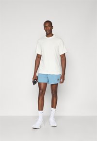Camiseta atlética de color gris claro, de mangas cortas, combinada con pantalones cortos de color azul claro que llevan un logo. El modelo sostiene una botella de agua negra y lleva zapatillas blancas.