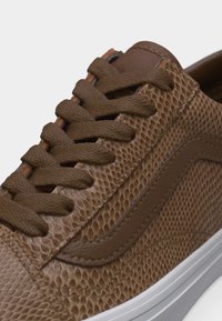 Καφέ αθλητικό παπούτσι με υφή φιδίσιο δέρμα, καφέ κορδόνια και λευκή σόλα, απεικονίζεται σε κοντινό πλάνο με έμφαση στην πλευρά και τα κορδόνια.