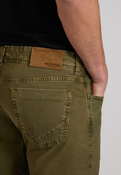 Close-up van de hand van een persoon in de voorzak van olijfgroene broek met een bruine lederen "Incotex" label op de achterkant van de tailleband.
