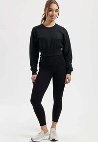 Schwarzer, kurz geschnittener Sweatshirt mit langen Ärmeln, kombiniert mit hoch taillierten schwarzen Leggings. Das Modell trägt hellgraue Sportschuhe.