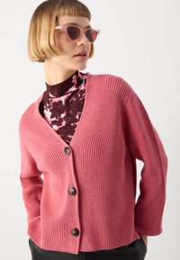 Rožinė raštuota kardiganas su V formos iškirpte, trimis sagomis ir ilgomis rankovėmis. Dėvimas ant tamsaus raštuoto marškinėlio; papildytas rožiniais akiniais nuo saulės.