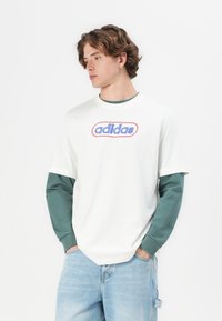Balta kokvilnas T-kreklu ar zilu un sarkanu "adidas" logo, virsū ar tumši zaļu garām piedurknēm kreklu, valkājas ar gaiši ziliem denima džinsiem.