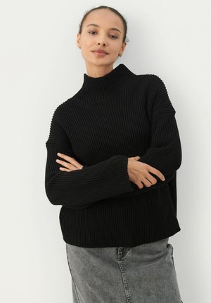 Schwarzer gerippter Pullover mit hohem Kragen, lockerer Passform und überschnittenen Schultern. Das Material wirkt weich und strukturiert, kombiniert mit einem grauen Jeansrock.