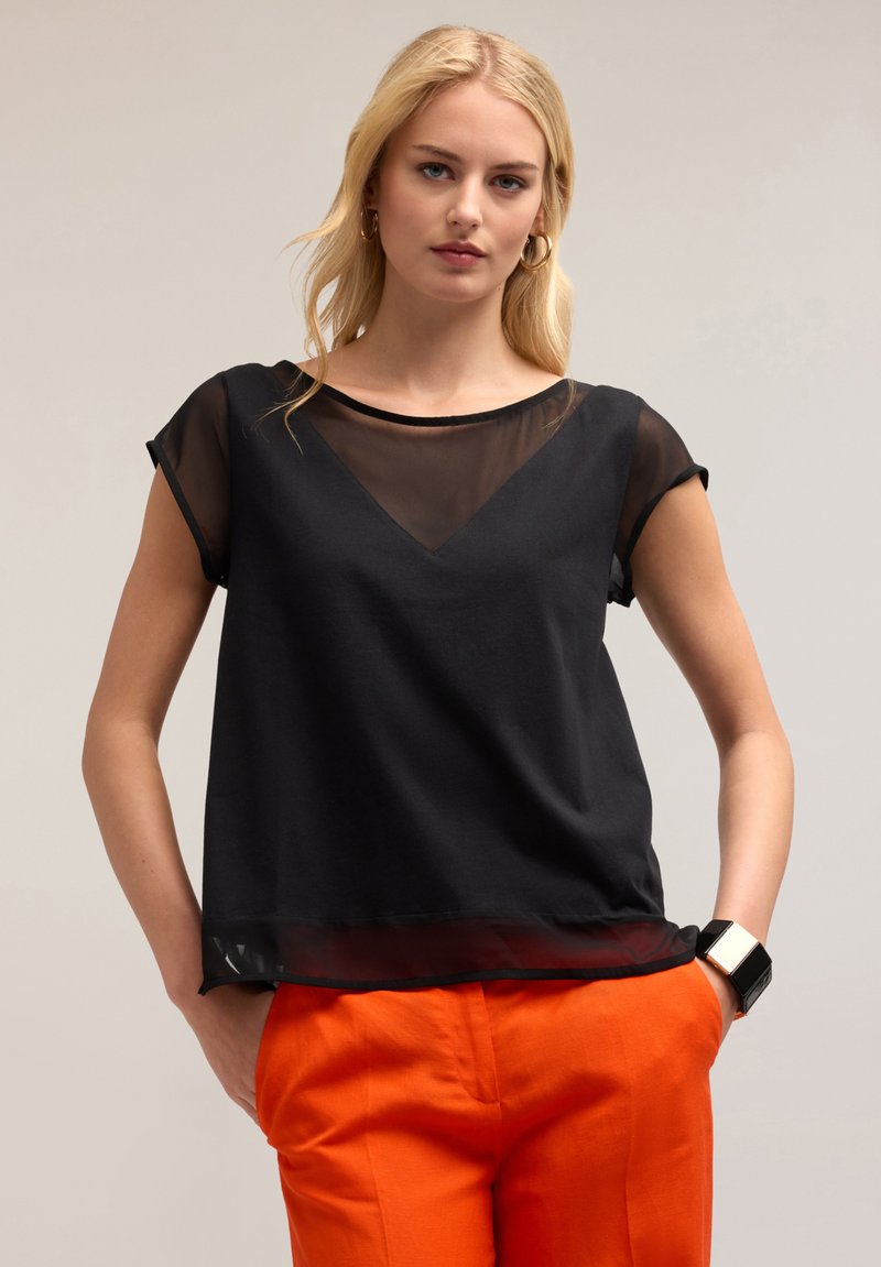 Zwarte top met een semi-transparante zwarte overlay, voorzien van korte mouwen en een V-vormige halslijn. Gekozen in combinatie met feloranje broek.