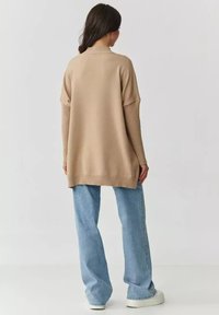 Beżowy, oversize sweter z prążkowanym golfem, miękka tekstura i długie rękawy, zestawiony z jasnoniebieskimi dżinsami o szerokich nogawkach i białymi trampkami.
