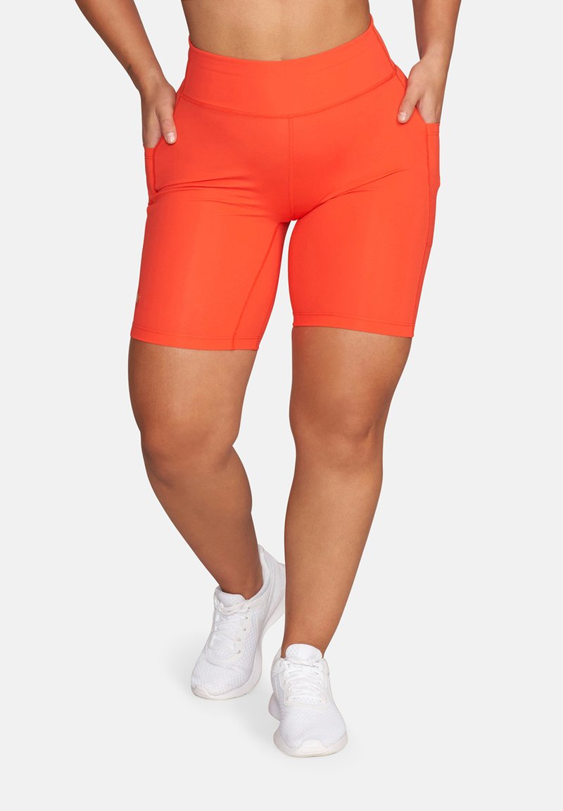 Person trägt leuchtend orangefarbene, hoch taillierte Biker-Shorts mit Taschen und weiße Sportschuhe, steht mit den Händen in den Taschen.