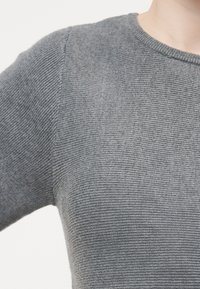 Κοντινό πλάνο ενός ατόμου που φοράει γκρι, ριγωτό πλεκτό πουλόβερ με μακριά μανίκια και στρογγυλή λαιμόκοψη, σε απλό φόντο.