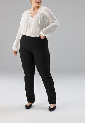 Weiße Langarmbluse mit gebundenem Ausschnitt, kombiniert mit schwarzen, eng anliegenden Hosen. Aus weichem Stoff mit klaren, maßgeschneiderten Kanten. Schwarze Ballerinas vervollständigen den Look.