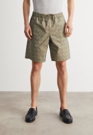 Grüne gemusterte Shorts mit elastischem Bund und Kordelzug, kombiniert mit schwarzen Loafers auf einem neutralen Bodenbelag.