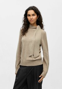 Pull en maille marron clair avec un col haut et un long d�étail de nouage à l'avant. Manches longues et base ajustée. Associé à un pantalon large noir.