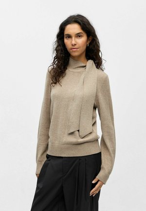 Pull en maille marron clair avec un col haut et un long détail de nouage à l'avant. Manches longues et base ajustée. Associé à un pantalon large noir.