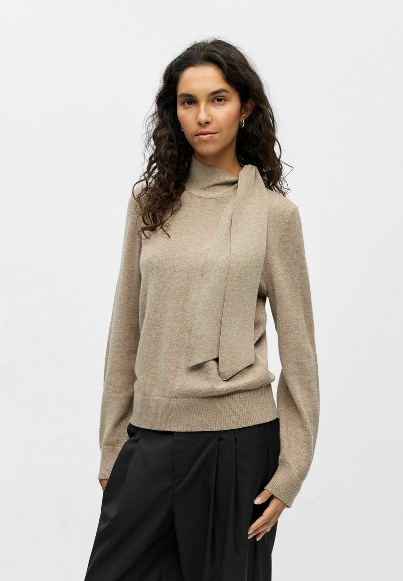 Pull en maille marron clair avec un col haut et un long détail de nouage à l'avant. Manches longues et base ajustée. Associé à un pantalon large noir.