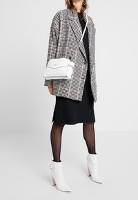 Donna che indossa un vestito nero, un blazer a motivo pied de poule, collant trasparenti a pois, stivali bianchi alla caviglia e una borsa a tracolla bianca.