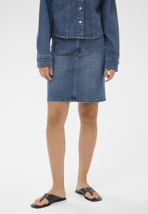 Kvinde iført en blå denimnederdel og matchende denimjakke, stående i sorte åbne flade sandaler mod en ensartet hvid baggrund.