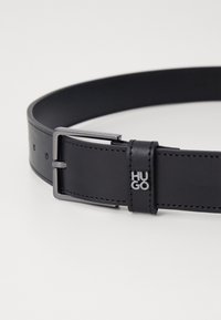 Svart läderbälte med en silverf�ärgad rektangulär spänne, med en liten "HUGO"-logotyp och synliga sömmar längs kanterna.