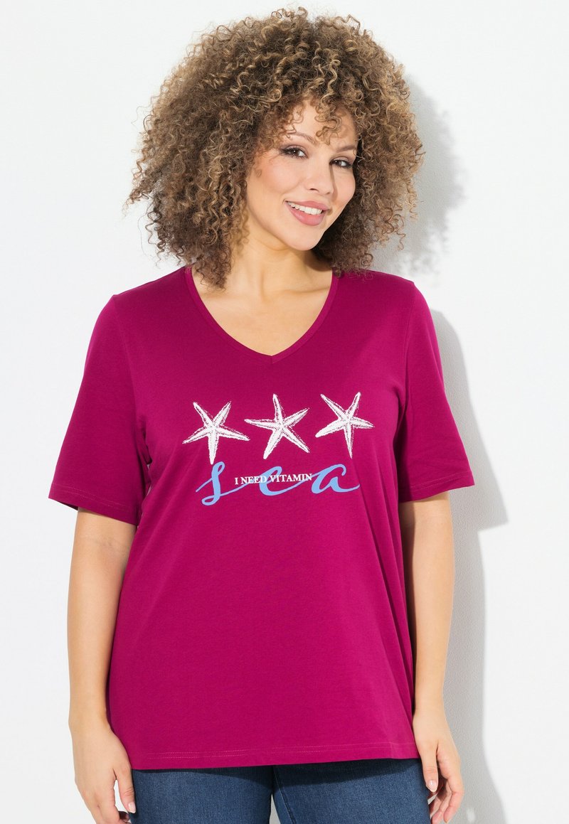 T-shirt fucsia con scollo a V e maniche corte, che presenta tre stelle marine e la scritta "Ho bisogno di vitamina mare" stampata in blu e bianco. Tessuto di cotone morbido.