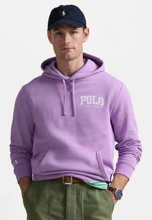 Fialová bavlněná mikina s kapucí a přední kapsou, s bílým logem "Polo Ralph Lauren"; nošena přes zelené kalhoty, doplněna námořnickou čepicí.
