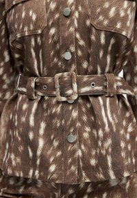 Braune Jeansjacke mit gepunktetem Muster, silbernen Druckknöpfen, doppelten Brusttaschen und einem breiten Stoffgürtel mit rechteckiger Schnalle.