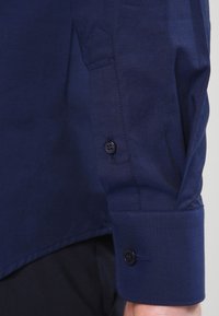 Mouwe blauwe lange mouw shirt manchet met twee donkere knopen, gedragen over zwarte broek, met zichtbare stofstructuur en stiksels.