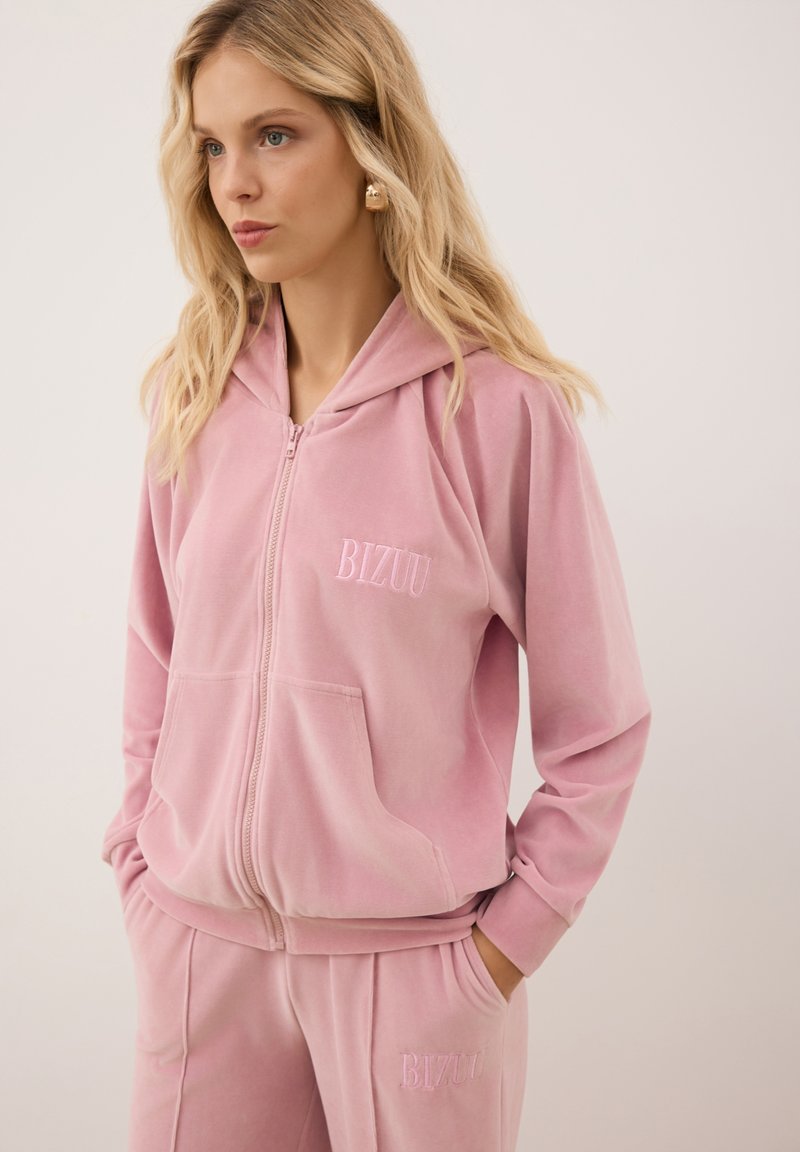 Roze rits hoodie van zacht materiaal, met een voorzak en geborduurd 'BIZUU' logo. In combinatie met bijpassende broek.