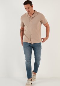 Beige kortærmet skjorte med knaplukning og krave, parret med tætsiddende blå jeans og lyse sneakers, model der går foran en neutral baggrund.