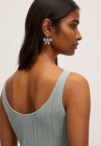 Lichtblauw geribde tanktop met een ronde halslijn, in combinatie met grote zilveren oorbellen in de vorm van bloemen. Zichtbaar detail van een rugtattoo.