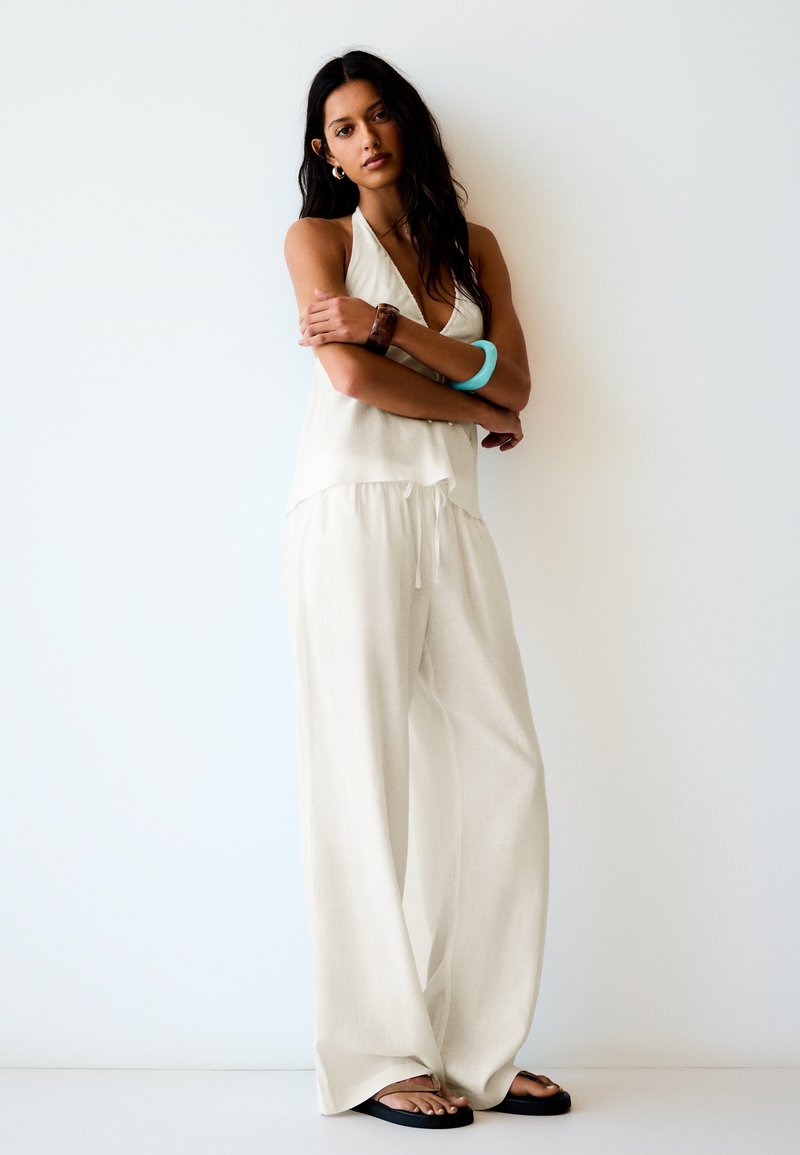 Femme aux longs cheveux foncés, portant un haut blanc sans manches à col halter et un pantalon blanc ample, avec deux bracelets, debout, bras croisés contre un mur blanc.