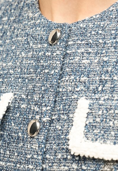 Μπλε και λευκό υφαντό ύφασμα με δύο λαμπερά ασημένια κουμπιά και πτερύγιο τσέπης με λευκή επένδυση σε ένα ένδυμα.