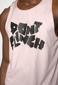 Persona con una camiseta sin mangas de malla rosa claro que lleva un texto negro en negrita que dice "NO TE TIRES" en el pecho.