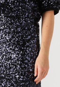 Close-up van een hand die naast een zwarte paillettenjurk met korte mouwen rust.