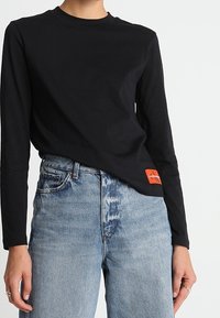 Chemise noire à manches longues en coton, avec un col rond et un patch rouge en bas à gauche avec du texte et un design blancs. Associée à un jean en denim bleu.