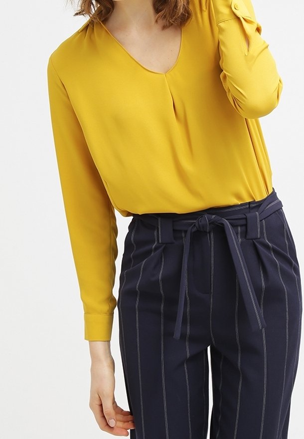 Blouse jaune moutarde en V avec des manches longues et des poignets boutonnés, associée à un pantalon taille haute à rayures navy avec une ceinture nouée.