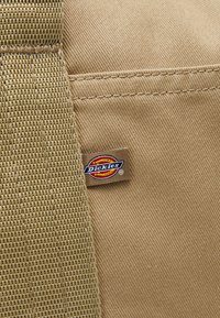 Vaalea kangaslaukkua, jossa on rakenne- ja sileitä alueita, ja sivussa pieni "Dickies"-logotägi. Ollaan näkyviä ompeleita.