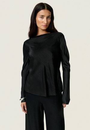 Žena s dlhými tmavými vlasmi, ktorá má na sebe lesklé čierne tričko s dlhým rukávom a čierne nohavice, stojaca pred jednoduchým svetlým pozadím.