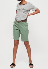 Camiseta gris con ribete negro y texto, combinada con pantalones cortos verde menta, un cinturón marrón y sandalias negras de deslizar. Atuendo casual de verano.