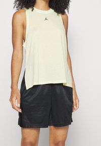 Top sin mangas de color amarillo claro con logo negro, hecho de tela transpirable, combinado con pantalones cortos de satén negros con aberturas laterales.