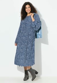 Kobieta z kręconymi włosami, ubrana w długą, niebieską sukienkę w wzory, szare botki za kostkę, z torbą na ramię z postarzanego denimu.