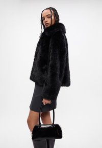 Casaco de pele sintética preto com gola alta, usado sobre uma mini saia cinza. Modelo a segurar uma mala patente preta; cabelo em tranças.