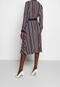 Pasiasta sukienka w kolorze granatowym, różowym i białym; długie rękawy, dopasowana talia i rozkloszowany dół. Noszona z białymi szpilkami.