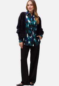 Cardigan nero fluffy a maniche lunghe sopra una camicia multicolore con bottoni, con motivi turchesi, blu e bianchi, abbinato a pantaloni neri e ballerine.