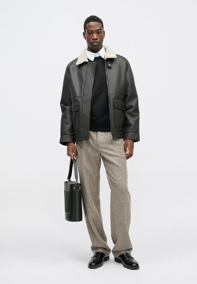 Čierna kožená bunda s krémovým ovčím golierom, skombinovaná s tmavým svetrom a béžovými károvanými nohavicami; drží čiernu štruktúrovanú tašku.
