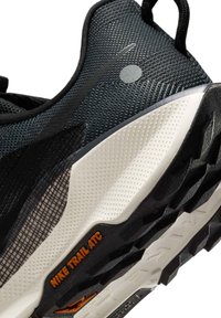 Zapatilla de trail running negra con un upper texturizado, que presenta un logo de Nike gris, una entresuela blanca y un detalle en naranja "NIKE TRAIL ATC" en la suela.