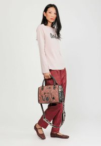 Borsa a tracolla rosa trapuntata con dettagli neri e testo decorativo, abbinata a una maglietta a maniche lunghe rosa chiaro e pantaloni bordeaux.
