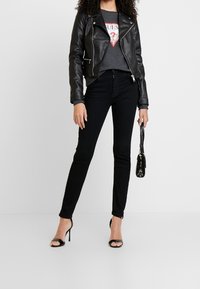 Svart läderbikerjacka över en grå t-shirt med tryck, kombinerad med svarta skinny jeans och svarta remklackar. Accessoarer inkluderar en liten svart väska.