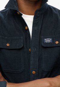 Granatowa koszula z korduroyu z dwiema kieszeniami na piersi, zapięciem na guziki oraz drewnianymi guzikami. Posiada etykietę na lewej kieszeni. Miękka w dotyku.