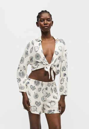 Femme portant une tenue blanche deux pièces avec des imprimés noirs de fruits et de feuilles, un crop top à manches longues noué, et un short assorti à taille élastique, debout.